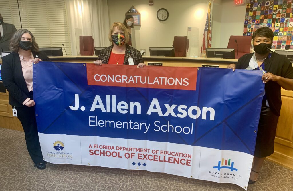 Superintendent, Board Member and school administrator hold banner Congratulations J. Allen Axson Elementary School a Florida Department of Education School of Excellence