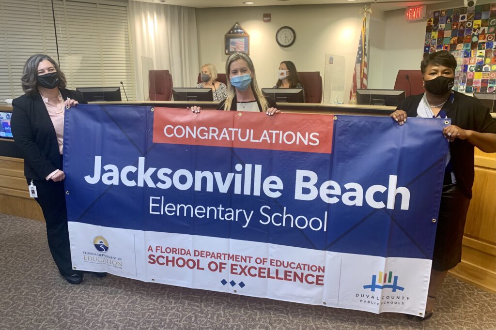 Superintendent, Board Member and school administrator hold banner Congratulations Jacksonville Beach Elementary School a Florida Department of Education School of Excellence