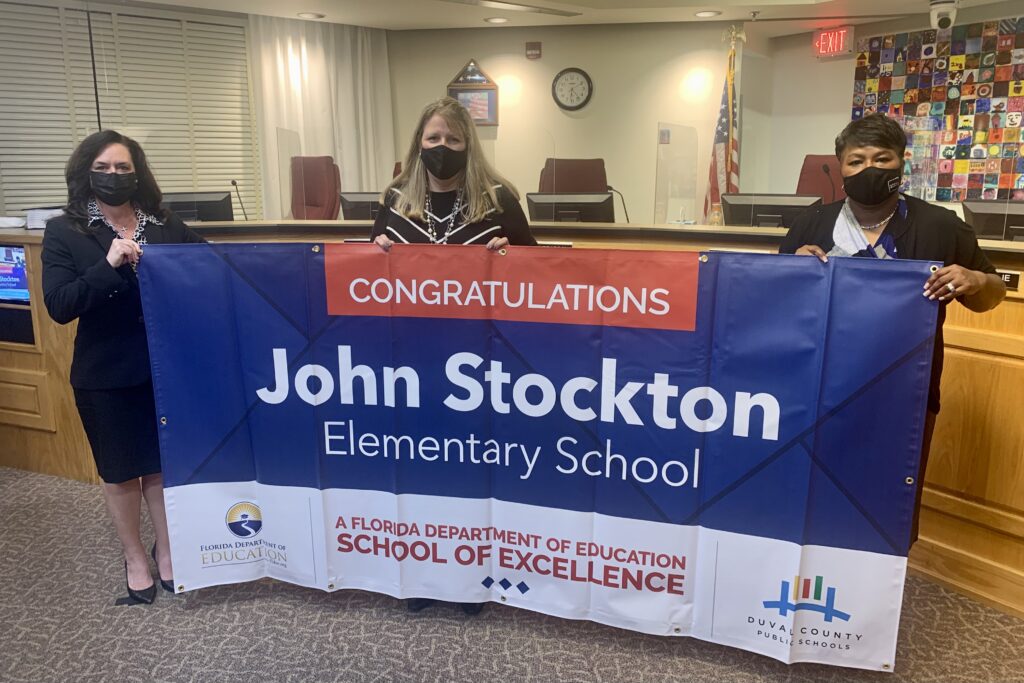 Superintendent, Board Member and school administrator hold banner Congratulations John Stockton Elementary School a Florida Department of Education School of Excellence