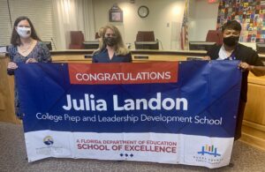 Superintendent, Board Member and school administrator hold banner Congratulations Julia Landon College Prep a Florida Department of Education School of Excellence