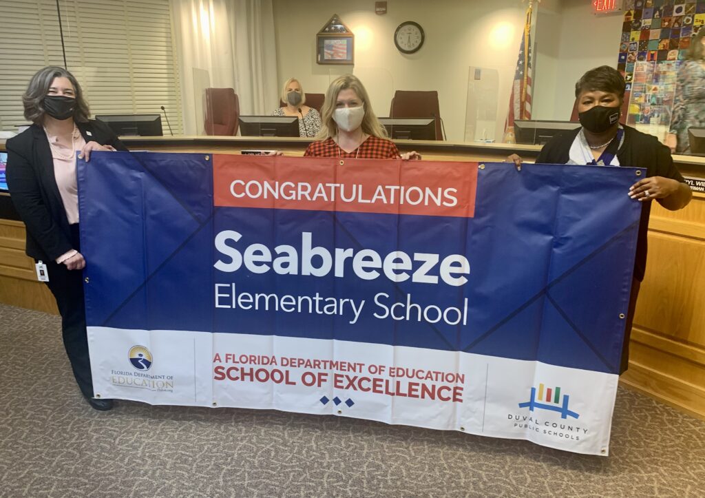 Superintendent, Board Member and school administrator hold banner Congratulations Seabreeze Elementary School a Florida Department of Education School of Excellence
